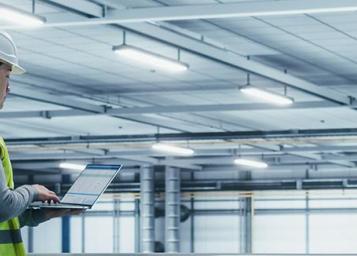 Man observes machinery with an ipad in hand, wearing a construction hat and vest on the left hand side of the image. 