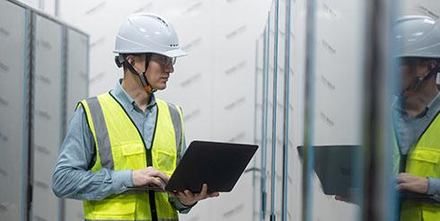 Man observes machinery with an ipad in hand, wearing a construction hat and vest on the left hand side of the image. 